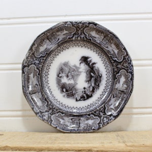 May include: A small, white porcelain plate with a black and white floral design. The plate has a scalloped edge and is decorated with a central image of a scene with trees and figures.