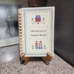 May include: A spiral-bound cookbook titled 6B Collection of Fabulous Recipes with a cream-colored cover featuring a blue house, a red heart, and two figures in blue and brown clothing, all displayed on a gold-colored plastic stand.