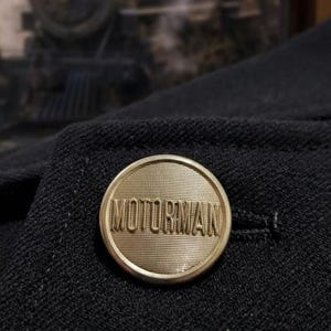 May include: Close-up of a black jacket with a gold-colored button. The button is round and embossed with the word "MOTORMAN". A vintage steam train is blurred in the background, suggesting a railway theme.