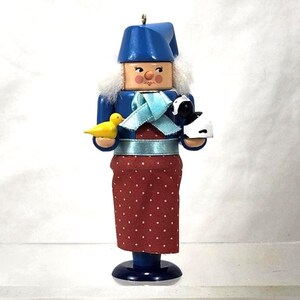 May include: A wooden nutcracker figurine in a blue hat and jacket, holding a yellow bird and a black and white sheep. The figurine has a red skirt with white polka dots and a light blue ribbon. The base is dark blue.