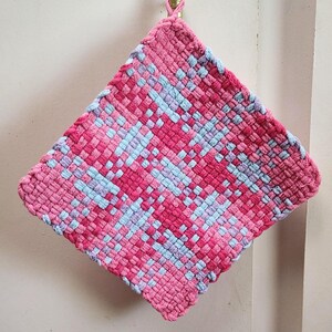 May include: A square, woven hot pad in shades of pink and light blue. The handmade pot holder is hanging from a gold hook. The woven design features a repeating pattern of pink and light blue squares.