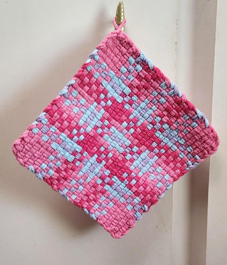 May include: A square, woven hot pad in shades of pink and light blue. The handmade pot holder is hanging from a gold hook. The woven design features a repeating pattern of pink and light blue squares.