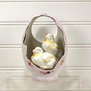 May include: A decorative, egg-shaped ceramic basket in a soft pink hue, adorned with delicate floral accents. Inside, two white ceramic birds with yellow and red details are nestled, creating a charming Easter or spring-themed display.