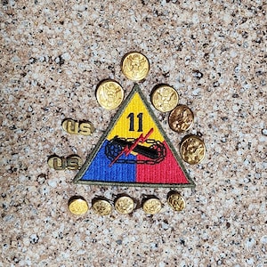 May include: A collection of military insignia, including a triangular patch with the number 11, a tank, and lightning bolts. Gold-colored buttons and two "US" pins are also present, all arranged on a textured surface.