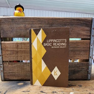 May include: A vintage copy of "Lippincott's Basic Reading" book with a brown cover and geometric design in yellow and white. A rubber duck wearing a black hat sits on a wooden crate in the background.