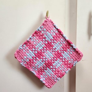 May include: A square, woven hot pad in shades of pink and light blue, hanging from a gold hook. The woven pattern creates a textured surface. The edges are finished with a pink border. This kitchen item is designed for heat resistance.
