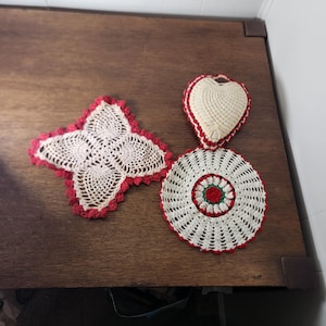 May include: A collection of handmade crochet items, including a four-point doily with red trim, a heart-shaped sachet, and a circular coaster with a red and green center. The items are displayed on a wooden surface.