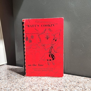 May include: A vintage cookbook titled "WATT'S COOKIN" with a red cover and black spiral binding. The cover features a cartoon chef character and the text "on the line". Compiled by the Rural Electrification Leaders of Jackson Electric Membership Corporation.