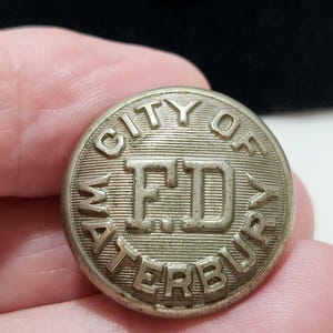 May include: A round, silver-toned metal button featuring a textured background with the embossed text CITY OF WATERBURY arranged in a circle and the letters F.D. prominently displayed in the center of the circular button face.