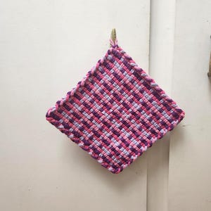 May include: A woven potholder in shades of pink, purple, and gray hangs against a white background. The square potholder has a braided edge and a loop for hanging. The woven design creates a textured appearance.