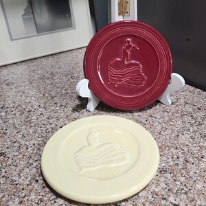 May include: Two decorative ceramic plates. One is a deep red with a raised design of a flamenco dancer. The other is a pale yellow with a similar dancer design. Both plates are round and have a ridged border.