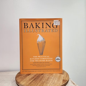 May include: An orange cookbook titled "BAKING ILLUSTRATED" with a whisk illustration. The book, "THE PRACTICAL KITCHEN COMPANION FOR THE HOME BAKER," sits on a wooden serving board. The book promises 350 recipes.