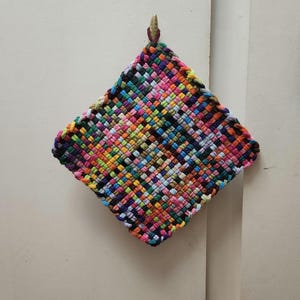 May include: A colorful, woven potholder in a diamond shape. The potholder features a vibrant mix of colors, including pink, green, yellow, and blue. It is hanging on a wall. The potholder is likely handmade.