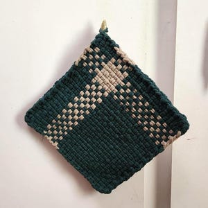 May include: A square, handmade macrame potholder in a dark green and tan woven pattern. The potholder is hanging on a white wall. The potholder is approximately 8 inches square.