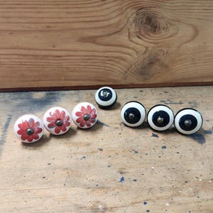 May include: A collection of decorative cabinet knobs. Three white knobs with red flower designs and three black and white knobs with brass-colored hardware are displayed. A single black and white knob is also present.