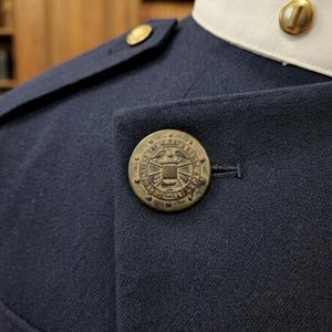 May include: Close-up of a navy blue uniform jacket with gold-colored buttons. The jacket features a white collar and a large, detailed button on the front. The buttons have a circular design with an emblem. The fabric appears to be a fine weave.