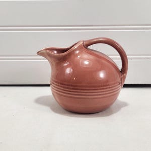 May include: A dusty rose ceramic creamer pitcher with a rounded, bulbous body and a smooth, glossy finish. The pitcher features a curved handle on the right side, a small pouring spout, and three horizontal embossed rings around the lower section.