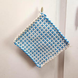 May include: Square woven potholder with a blue and white checkered pattern. The edges are trimmed with a blue border. A small gold hook is attached to the top corner for hanging. The potholder is designed for kitchen use.