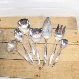 May include: A set of eight silver-plated serving utensils, including a ladle, a serving spoon, a slotted spoon, a fork, a butter knife, and two spoons. The utensils are arranged on a wooden surface.