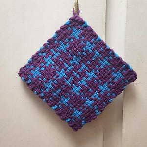 May include: A square, woven potholder in a diamond pattern. The potholder is primarily purple with a repeating pattern of light blue squares. A loop for hanging is at the top. The potholder is approximately 8 inches square.
