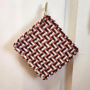 May include: A square, woven hot pad with a geometric pattern. The design features alternating bands of white, rust, and burgundy. A loop of white cord is attached for hanging. The hot pad is hanging on a gold-colored hook.