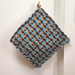 May include: A square, woven potholder in a diamond pattern. The potholder features a repeating pattern of blue, tan, and burgundy threads. A small gold-colored loop is attached to the top corner for hanging. Kitchen and home decor.