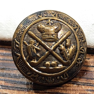 May include: A round, antique-style brass button featuring a central embossed design of a crown above two crossed golf clubs. The emblem is flanked by a small figure and a thistle, with a textured border containing decorative lettering and symbols.