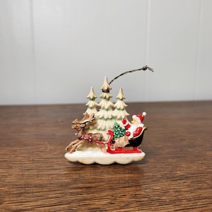 May include: A Christmas ornament featuring Santa Claus in a sleigh pulled by a reindeer, with a backdrop of snow-covered pine trees. The ornament is painted with red, white, green, and brown colors, and is set on a wooden surface.