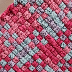 May include: Close-up of a woven textile in a square pattern. The textile features a mix of pink, red, light blue, and purple threads. The weave creates a textured surface, with the colors alternating in a grid-like design. The edges are finished with a pink border.