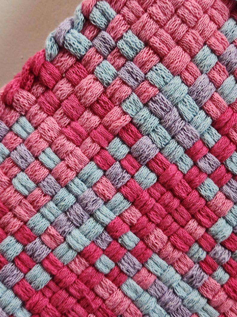 May include: Close-up of a woven textile in a square pattern. The textile features a mix of pink, red, light blue, and purple threads. The weave creates a textured surface, with the colors alternating in a grid-like design. The edges are finished with a pink border.
