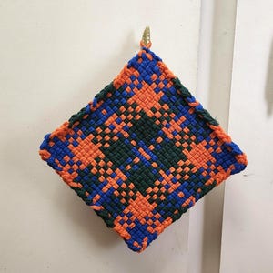 May include: A square, woven hot pad in a diamond pattern. The design features orange, blue, and green colors. The edges are finished with a blue border. A small, gold-colored loop is attached to the top corner for hanging.
