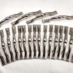 May include: A collection of silver-toned cabinet hardware, including handles and pulls. The handles have a twisted design, while the pulls feature a rectangular top and a decorative, rounded base. The hardware is arranged on a white surface.