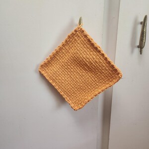 May include: An orange square woven potholder hanging on a gold hook. The potholder has a textured surface and a decorative edge. The background is a plain white wall and a white door with a gold handle.