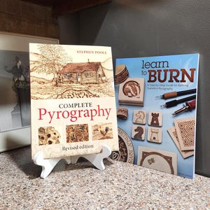 May include: Two books on a white stand. The book on the left is titled "Complete Pyrography" with a cabin illustration. The book on the right is titled "Learn to Burn" with pyrography tools and examples. Both books are on a light-colored surface.