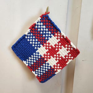 May include: A square, woven hot pad in a patriotic color scheme. The design features a blue corner, a red corner, and a white and red checkered pattern. A small loop is attached for hanging.