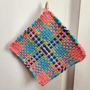 May include: A square, woven hot pad in a diamond pattern. The pad features a color scheme of coral, blue, green, and purple. A small, gold-colored loop is attached to the top corner for hanging. The woven design is intricate and detailed.
