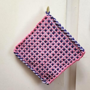 May include: A square, woven hot pad in a pink, purple, and gray checkered pattern. The edges are trimmed with a braided purple border. A small gold loop is attached for hanging. Kitchen decor.