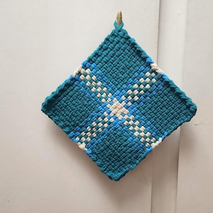May include: A square, woven potholder in shades of teal, blue, and white. The design features a central cross pattern with a gold-colored loop for hanging. The potholder is made of a tightly woven material, suitable for kitchen use.