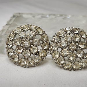 May include: A pair of circular silver-toned earrings featuring a cluster of clear, faceted round rhinestones set in a lattice pattern. Each earring is composed of multiple small, sparkling stones arranged in a dense, circular formation.