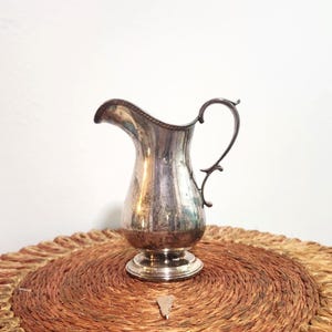 May include: A silver-colored pitcher with a decorative handle and spout. The pitcher has a bulbous body and sits on a round base. The pitcher is placed on a round, woven mat.