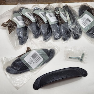 May include: A collection of black cabinet pulls, some still in their original packaging. The pulls have a hammered texture and a curved design. Also included are screws for installation. The hardware is suitable for furniture or cabinetry.