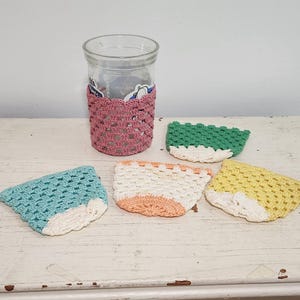 May include: A clear glass jar with a pink crocheted cozy and four crocheted coasters in blue, green, white, and yellow. The coasters have a white border. The items are on a white, distressed wooden surface.