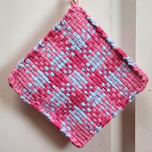 May include: A square, woven hot pad in shades of pink and light blue. The handmade pot holder is hanging from a gold-colored hook. The woven design creates a textured surface, perfect for kitchen use.