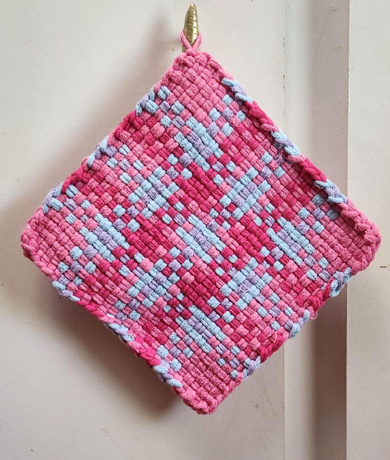 May include: A square, woven hot pad in shades of pink and light blue. The handmade pot holder is hanging from a gold-colored hook. The woven design creates a textured surface, perfect for kitchen use.