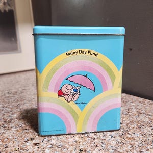 May include: A rectangular, light blue metal tin with a design featuring a rainbow and a cartoon character under a pink umbrella. The text "Rainy Day Fund" is arched above the rainbow. The tin is on a speckled countertop.
