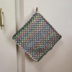May include: A square, woven potholder in a diamond pattern. The potholder is primarily gray, with accents of pink, green, and blue woven throughout. It hangs from a brass hook on a white surface.