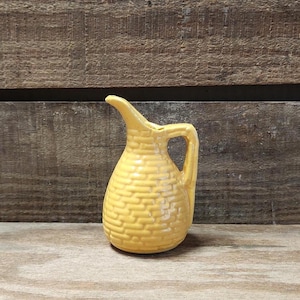 May include: A small, yellow ceramic pitcher with a textured, basketweave design. The pitcher has a curved spout and a handle. The background is a wooden surface and a wooden wall.