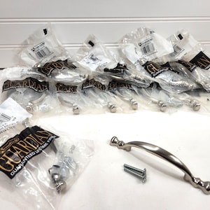 May include: A collection of new cabinet pulls in clear plastic packaging. The pulls are silver-toned with a curved design and decorative ends. The packaging includes the brand name "Guardian" and the phrase "Guaranteed Lifetime Finish".