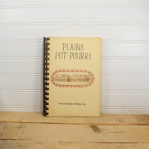 May include: A vintage cookbook titled "Plains Pot Pourri" with a spiral binding. The cover is beige with the title in brown text and an illustration of a town. The text "Food Favorites of Plains, Ga." is also visible.