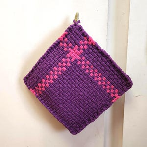 May include: A square, handmade purple potholder with a woven pink cross design. The potholder is hanging on a wall. The potholder is made of a thick, textured material.